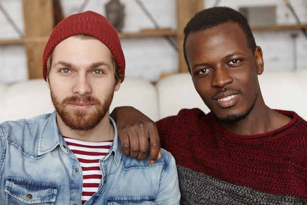 People, love and homosexuality. Samesex male couple spending nice time together at cafe: Afro-American man holding hand on his Caucasian partner's shoulder, both looking and smiling