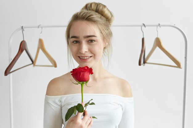 People, love, romance, beauty and affection concept. Attractive young Caucasian woman wearing white open shoulders top, smiling, holding one red rose from her unknown secret admirer