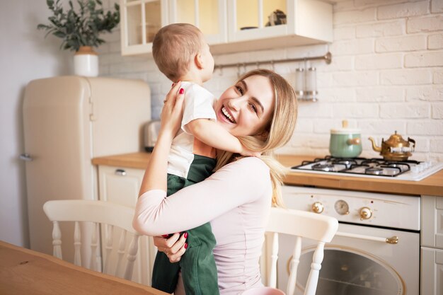 People, maternity, love, family and relationships concept. Portrait of happy pretty young woman sitting in stylish kitchen interior hugging her adorable baby son, looking with joyful smile