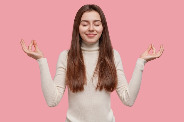 People and meditation concept. Relaxed peaceful young woman holds hands in mudra sign, keeps eyes closed, relaxes after work in office