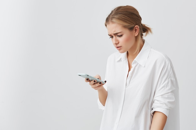 Free photo people and modern communication. studio portrait of worried pretty blonde girl reading urgent text message via smart phone. young female looking confused holding cell phone in her hand.