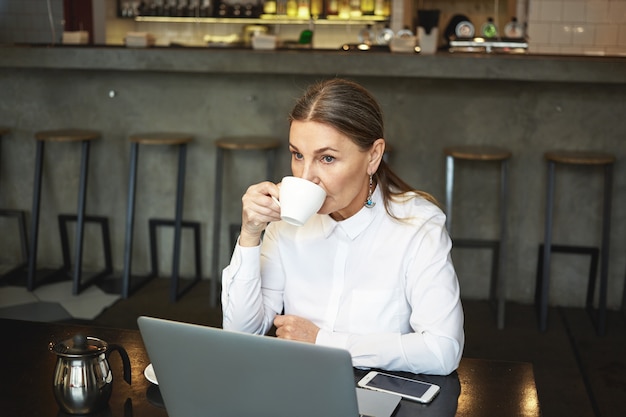 People, modern lifestyle, technologies, communication and leisure concept. Serious thoughtful retired lady with gray hair using laptop pc for remote work while having coffee at cafeteria alone