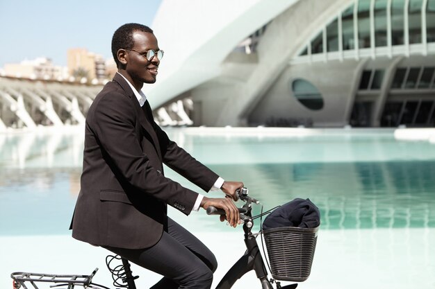 People, modern lifestyle, transport and ecology concept. Successful ecologically friendly dark-skinned male head of large financial company going to office on bicycle, wearing black formal suit