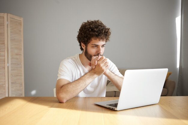 People, modern technology, communication, job and occupation concept. Stylish student with curly hair and beard sitting in front of open laptop at wooden desk, reading scientific article online