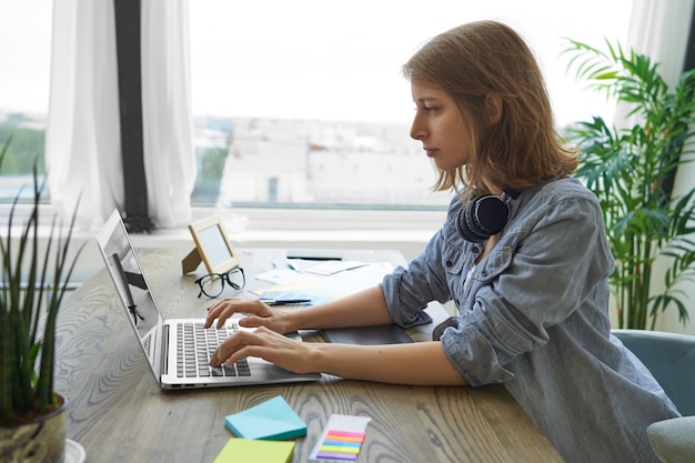 사람, 현대 기술 및 직업 개념. 나무 책상에 창 옆에 앉아 휴대용 컴퓨터에 그녀의 목 keyboarding 주위에 헤드폰 심각한 집중된 젊은 여성 프리랜서의 측면보기