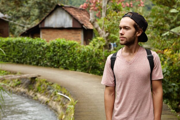 People, nature and summer concept. Stylish young unshaven hipster man wearing snapback and backpack relaxing outdoors while walking along country road in rural area