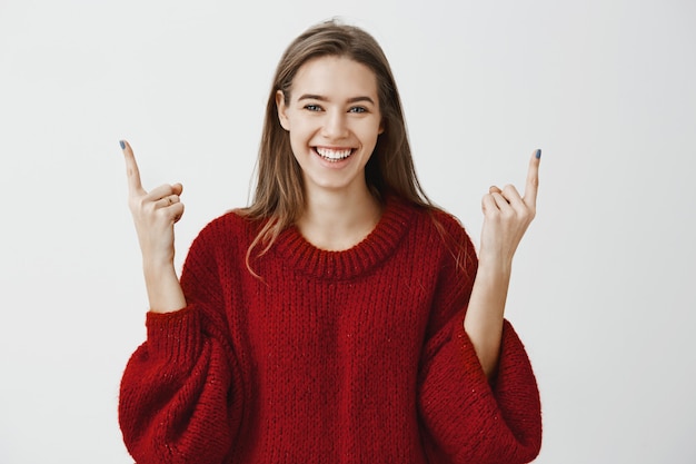 People need to lift heads up and live on bright side. satisfied beautiful female student in trendy loose sweater, raising index fingers and pointing up with broad pleased smile over gray wall