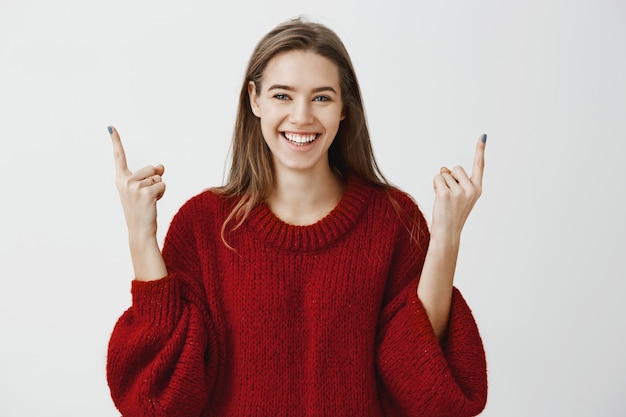 People need to lift heads up and live on bright side. Satisfied beautiful female student in trendy loose sweater, raising index fingers and pointing up with broad pleased smile over gray wall