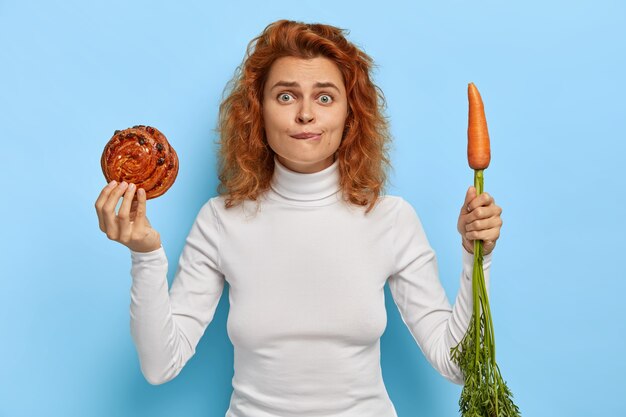 Люди, питание, диета и концепция нездоровой пищи. Смущенная рыжая женщина держит свежую вкусную булочку и морковь, выбирает между овощами и кондитерскими изделиями, носит белую водолазку, стоит в помещении