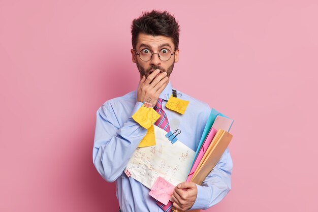 People and occupation concept. Shocked man manager in round spectacles covers mouth and stares surprised at camera holds documentation prepares for business meeting 