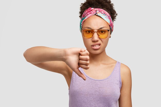 People and pessimism concept. Discontent African American female with negative facial expression, shows dislike gesture, doesn`t like something, dressed in casual purple t-shirt, isolated on white