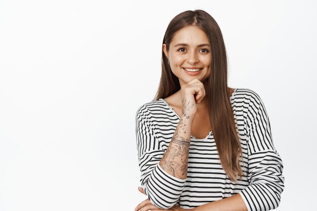 People portraits. Young woman smiling, looking with interest at camera, thinking of smth, hold hand on chin, standing against white background.