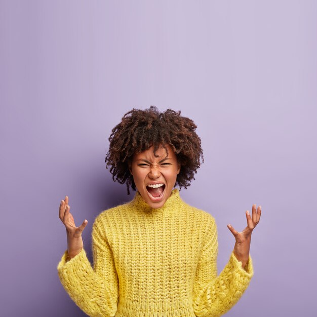People, rage, irritation concept. Emotional outraged crazy woman gestures angrily, raises hands, shouts madly on husband, expresses negative emotions, wears yellow clothes, isolated on purple wall.