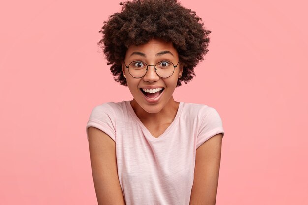 People and reaction concept. Happy overjoyed young African American female reacts on positive news, has broad smile and surprised expression, wears casual t-shirt, poses against pink wall