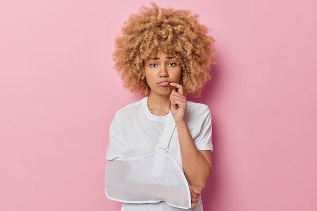 Foto gratuita concetto di recupero e riabilitazione delle persone la donna europea dai capelli ricci insoddisfatta guarda infelicemente alla telecamera che ha rotto il braccio dopo l'incidente indossa una maglietta bianca isolata su sfondo rosa