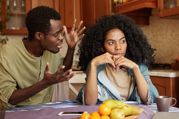 人との関係の概念。キッチンで主張しているアフリカ系アメリカ人のカップル：完全に彼を無視している彼の美しい不幸なガールフレンドに叫んで、怒りと絶望の中で身振りで示す眼鏡の男