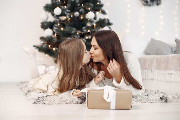 People reparing for Christmas. Mother playing with her daughter. Family sitting by the Christmass tree. Little girl in a white dress.