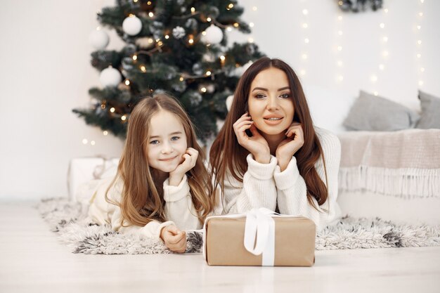 People reparing for Christmas. Mother playing with her daughter. Family sitting by the Christmass tree. Little girl in a white dress.