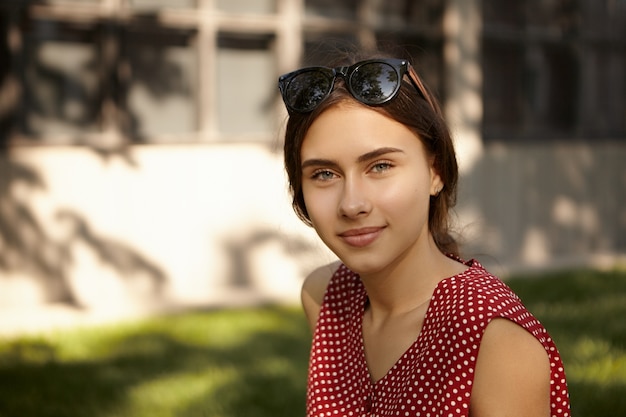 Persone, riposo, bellezza, stile e concetto di moda. ritratto di carino affascinante giovane donna caucasica con sfumature sulla sua testa godendo una bella giornata estiva all'aperto