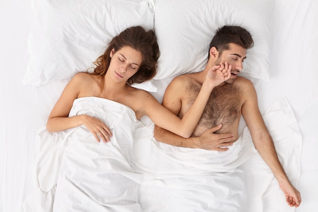 人々、休息と睡眠の概念。リラックスした家族のカップルは快適なベッドで安らかに眠り、楽しい夢を見て、女性は夫に手を伸ばし、怠惰な一日を過ごし、非常に早く目覚めたくありません。