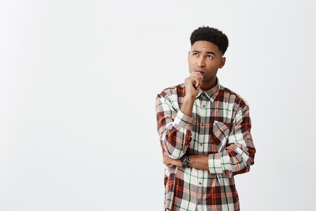 Free photo people's emotions. close up portrait of young attractive dark-skinned men with curly hair in casual shirt holding hand near mouth with thoughtful expression, being curious about film he saw.