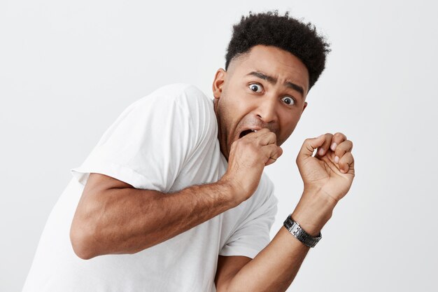 People's emotions. Close up of young handsome dark-skinned american guy with afro hairdo in white t-shirt holding hands near face with frightened expression, watching horror movie at night.