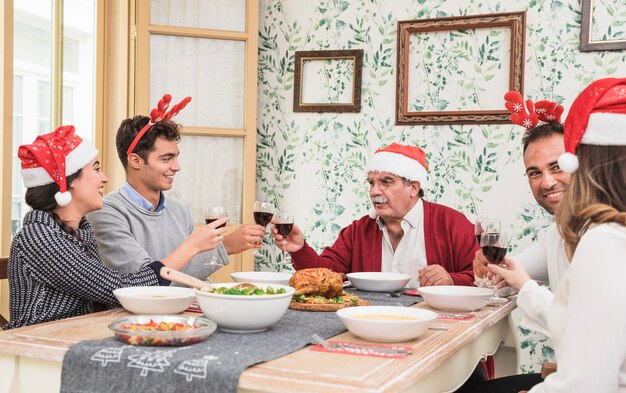 サンタの帽子の人々は、お祝いのテーブルで眼鏡をつまむ