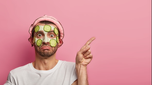 People, skin care and beauty concept. Surprised young man applies facial mask with fresh cucumbers, points index finger on blank space