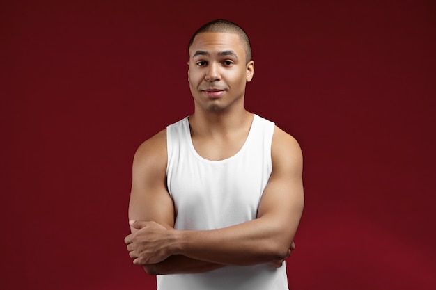 People, sports, fitness and active healthy lifestyle concept. Attractive fit young mixed race male with muscular shoulders posing in studio, keeping arms folded, his look expressing confidence