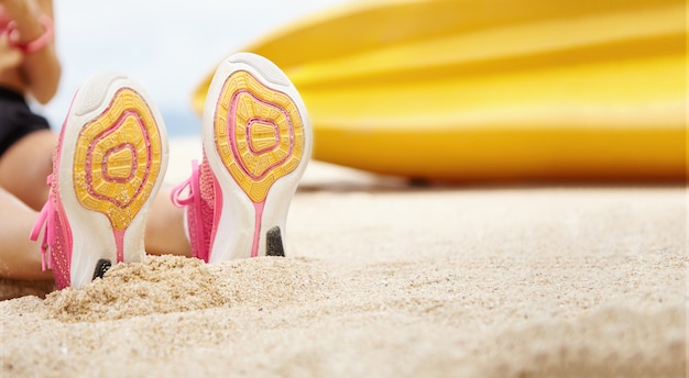 Foto gratuita persone, sport e stile di vita sano. chiuda sulla vista delle suole delle scarpe da corsa femminili. giovane sportiva che ha resto fuori, seduto sulla spiaggia dopo un intenso esercizio cardio. morte superficiale di campo