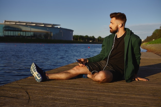Free photo people, sports, modern lifestyle and technology concept. portrait of fashionable young bearded man wearing stylish clothes relaxing by lake in cityscape, listening to audiobook or music tracks