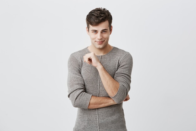 People, style, fashion concept. Handsome young European male with stylish haircut and blue eyes, wearing sweater posing indoors, keeping arms folded, looking  with handsome flirting smile