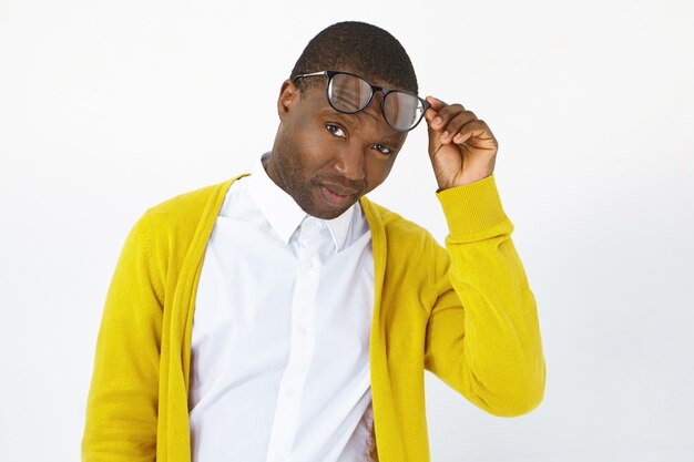 People, style, fashion, optics, eyewear and vision concept. Picture of stylish young dark skinned man posing in studio, raising trendy eyes glasses and staring at camera with sceptic expression