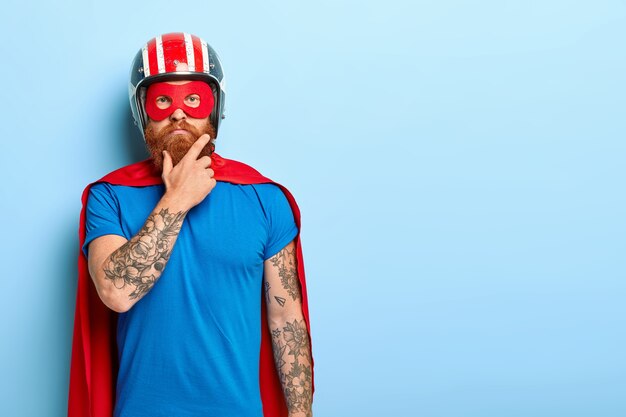 人と超大国のコンセプト。赤い太いひげを持つ真面目な男は、ヘルメットと赤いスーパーヒーローのマントを着ています
