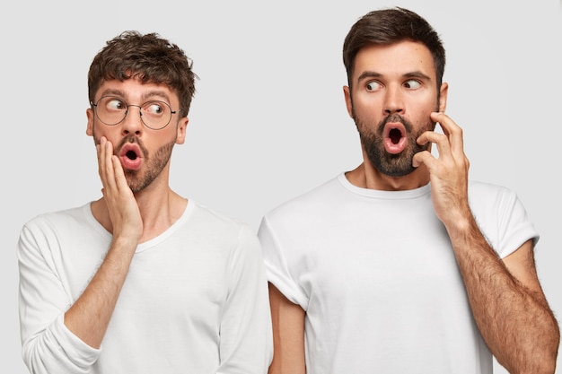 People and surprisement concept. Stunned bearded young men with widely opened mouthes and bugged eyes, look with shocked expressions at each other, hear unexpected news, isolated over white wall