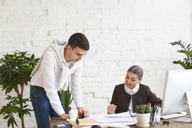 People, teamwork, cooperation and job concept. attractive young male architect standing at desk holding pencil while showing technical drawings to his middle aged female boss in modern office interior