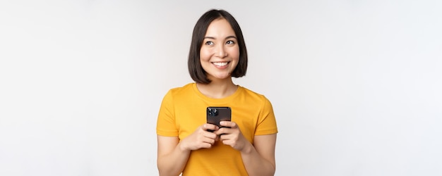 People and technology concept Smiling asian girl using smartphone texting on mobile phone standing against white background