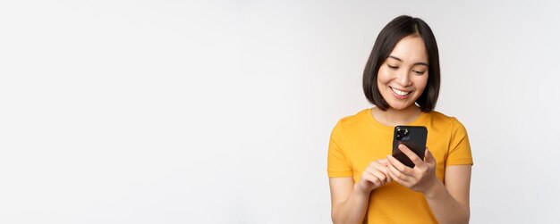 People and technology concept smiling asian girl using smartphone texting on mobile phone standing a