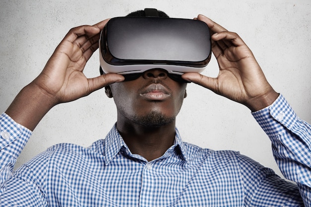 People, technology, cyberspace and entertainment concept. African man dressed in checkered shirt using 3d headset, playing video games.