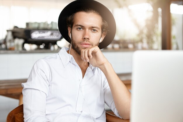 People, technology and leisure concept. Fashionable young man with beard listening to songs on earphones, using online music app on his laptop computer