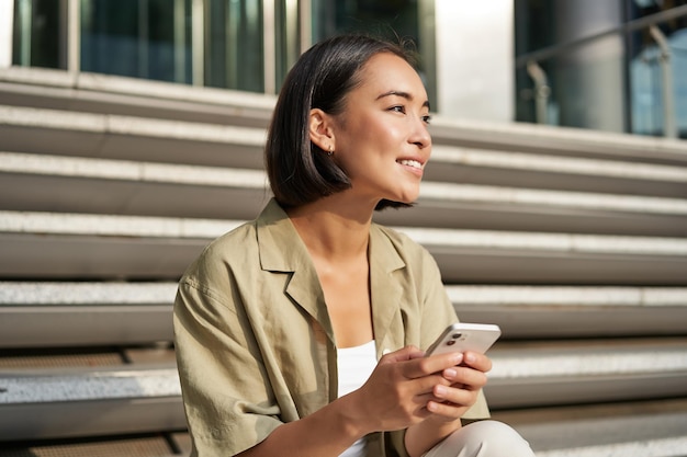 Free photo people and technology smiling beautiful asian woman sitting on stairs in city holding mobile phone g