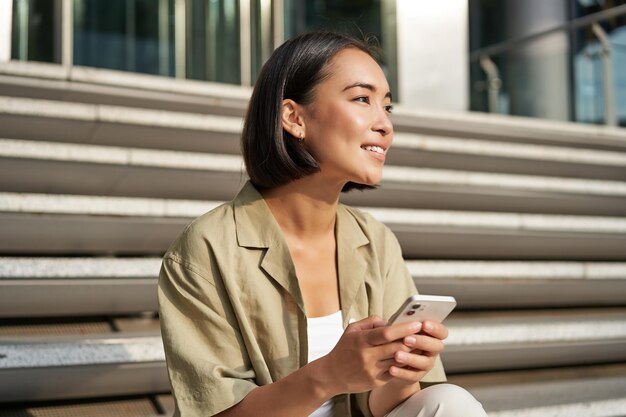 People and technology smiling beautiful asian woman sitting on stairs in city holding mobile phone g