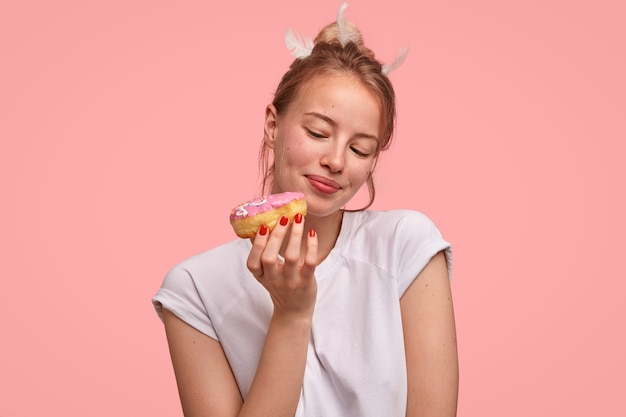 People and temptation concept. Attractive young European female looks at sweet doughnut, going to eat for breakfast, fonds of junk food, dressed casually, isolated over pink wall