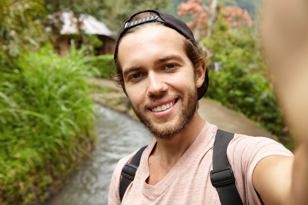 People, travel and adventure concept. Attractive young bearded adventurer wearing backpack and cap taking selfie