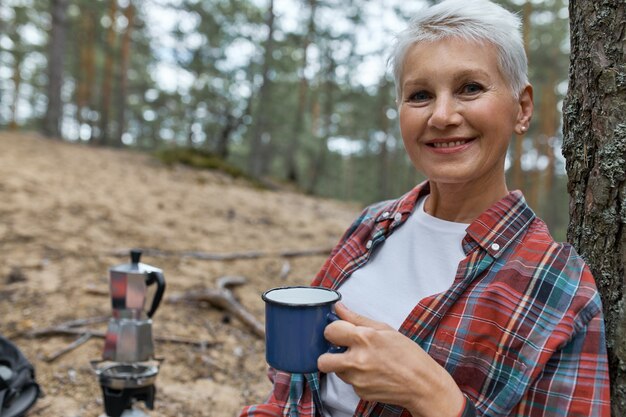 People, travel, hiking and vacations. Joyful middle aged Caucasian woman posing outdoors with cup, drinking tea in wild nature, having rest on camping site in pine forest, enjoying peaceful atmosphere