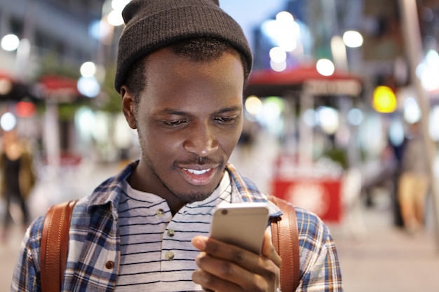 People, travel, lifestyle and modern technology. Headshot of positive young Afro American backpacker using online navigation app on his smartphone while walking around foreign city in the evening