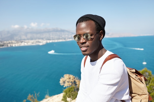 People, travel, tourism and lifestyle. Stylish dark-skinned traveler wearing trendy clothing standing on mountain above sea