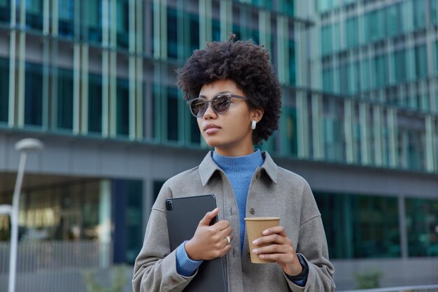 People and urban lifestyle concept Stylish curly haired woman walks outdoors with digital tablet and takeaway coffee focused aside wears sunglasses and jacket stands against modern glass building