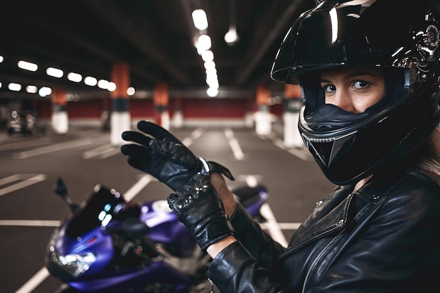 People, urban lifestyle, extreme sports and adrenaline conept. Sideways portrait of palyful styligh young Caucasian motorcycle rider in fashionable black leather jacket and helmet, adjusting gloves