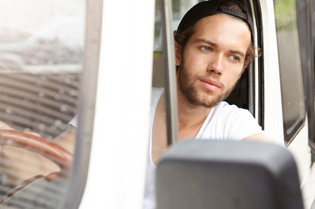 People, vacations and travel concept. Handsome young bearded man sitting inside his white SUV vehicle and looking out of open window with joyful expression